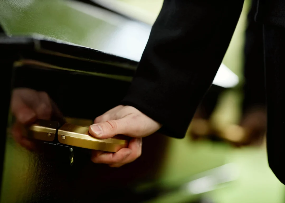 A person wearing a black suit holds the gold handle of a black coffin.